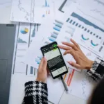 Pebisnis menganalisis data keuangan di smartphone dengan laptop dan dokumen